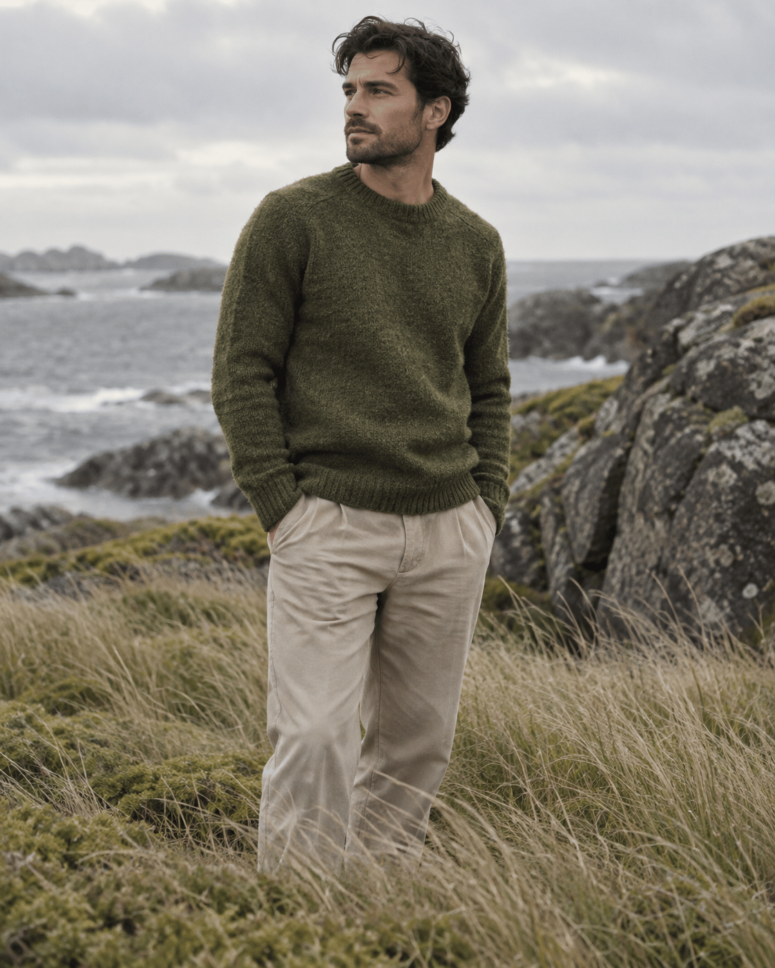 Man wearing a moss green knit and beige trousers on a rocky coast